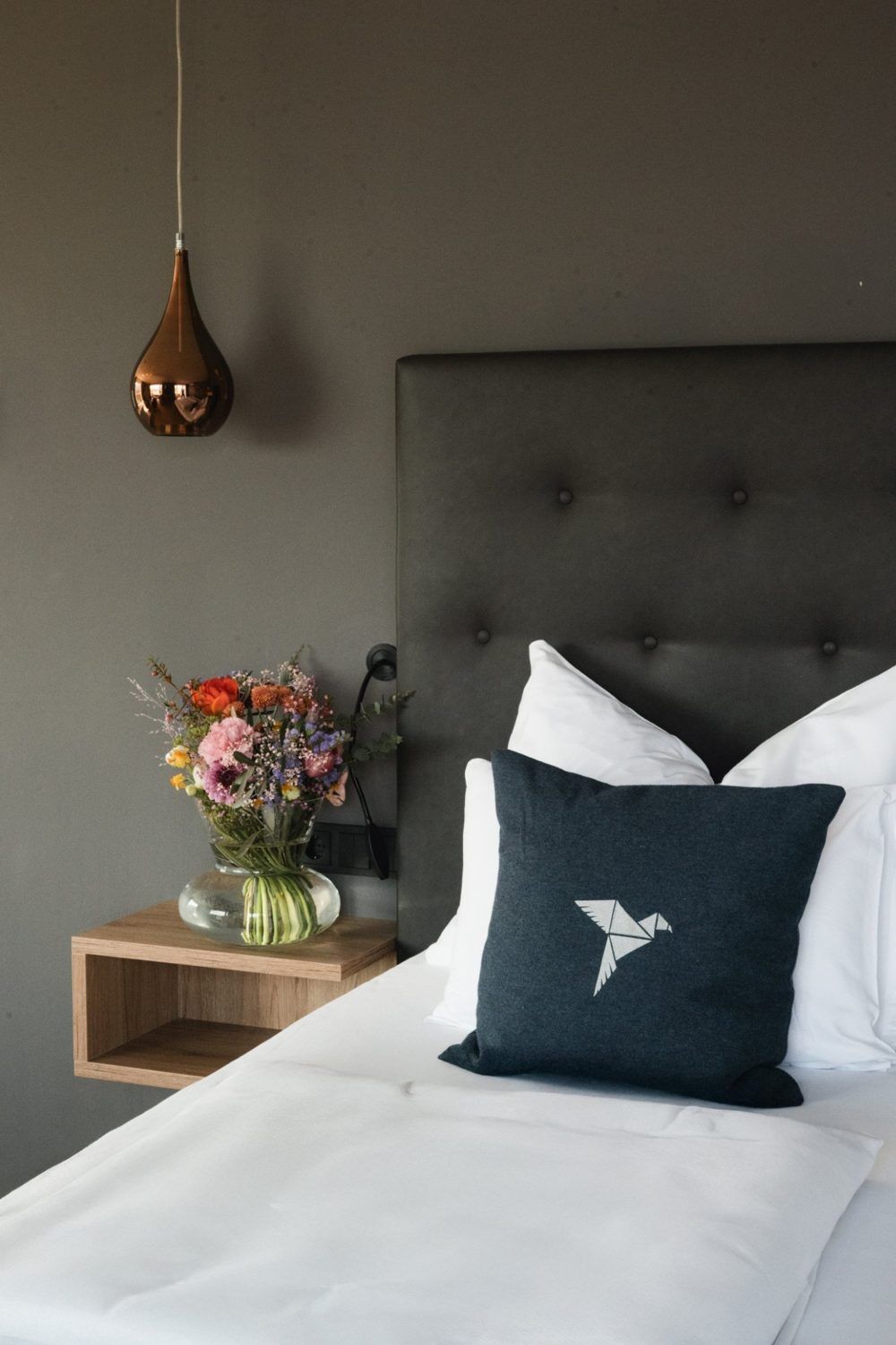 Hotelzimmer im das MEI: Bett mit dunkelblauem Kissen mit Vogel-Logo, Nachttisch mit Blumen und Hängeleuchte