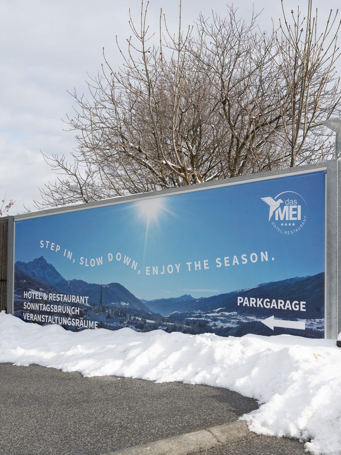 Werbebanner von das MEI „Step in, slow down, enjoy the season“ an einer Einfahrt im Schnee, vor Bergpanorama