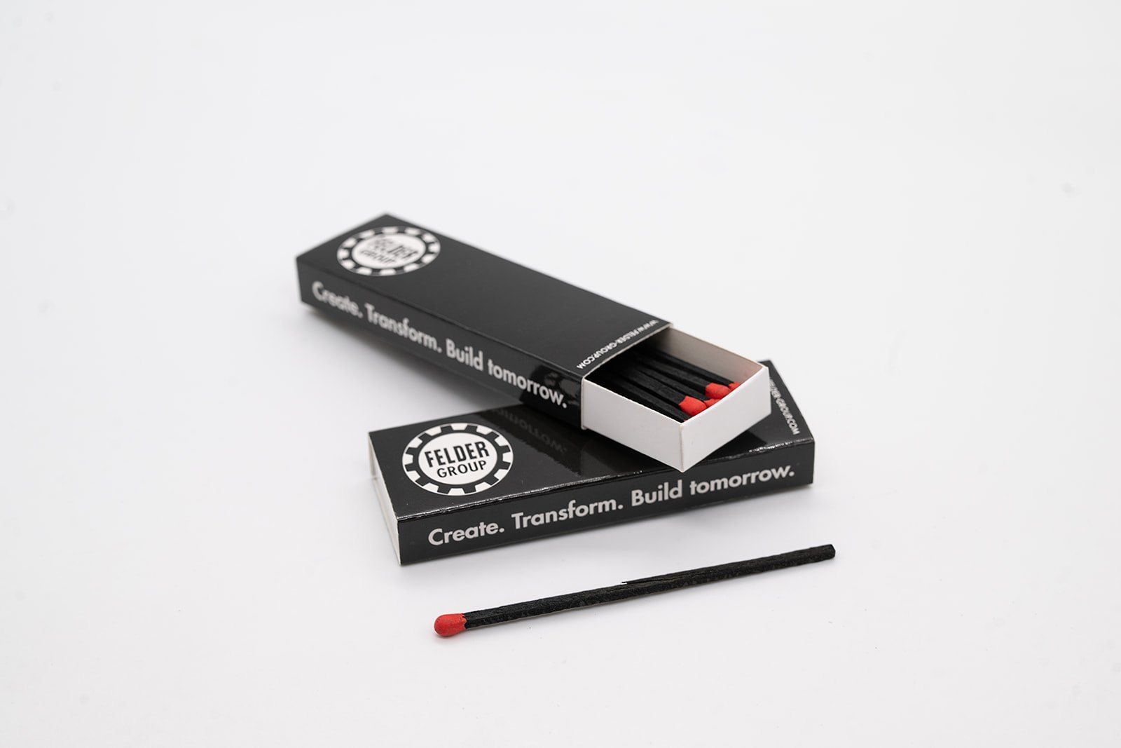 Geöffnete schwarze Zündholzschachtel mit Felder-Logo und ein Zündholz auf weißem Grund